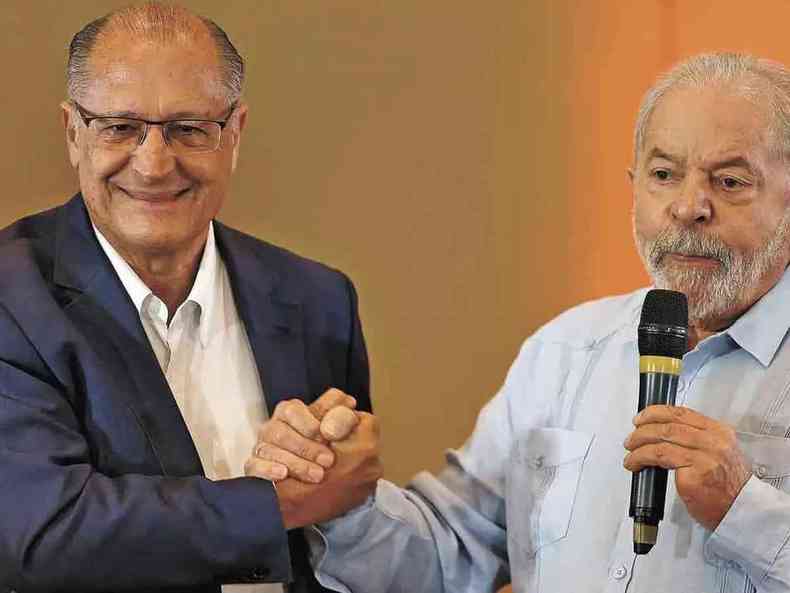 Lula e Alckimin dão as maõs em gesto de parceria. Lula está discursando com o microfone na outra mão.
