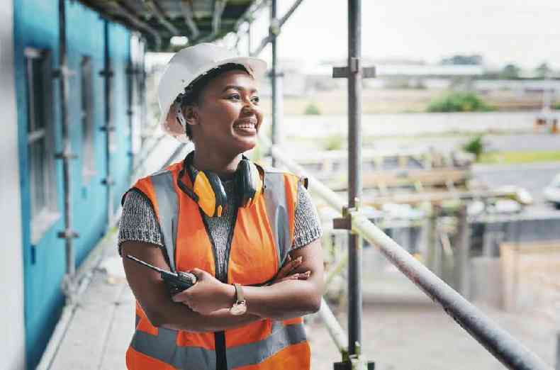 Foto colorida mostra trabalhadora negra com uniforme em uma construo, sorrindo