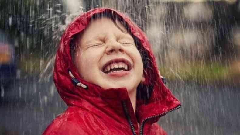 Criança de casaco vermelho com capuz sorrindo enquanto toma chuva 
