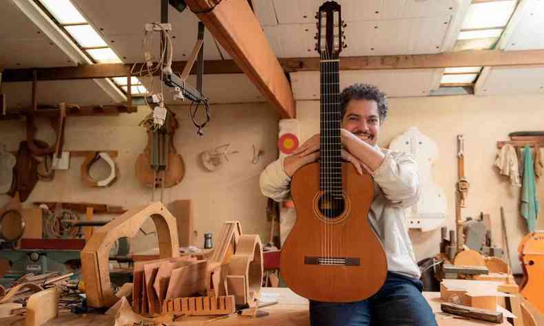 de cala jeans e camisa branca, Thiago Delegado segura violo no colo, sentado em mesa de marcenaria com instrumentos sendo construdos