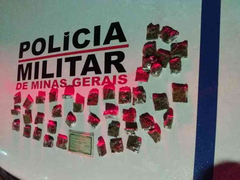 70 embalagens com maconha apreendidas pela PM e uma identidade falsa. Os objetos estão sobre uma mesa com o logotipo da Polícia Militar de Minas Gerais.