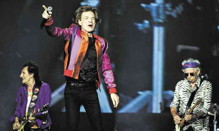 Mick Jagger segura microfone e aponta para a plateia no palco, com Ronnie Wood e Keith Richards atrs de si