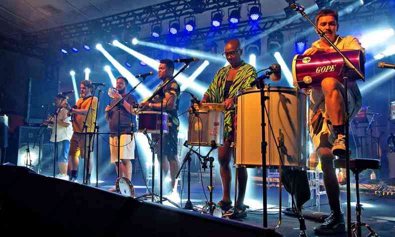 Seis integrantes do bloco Samba do Queixinho se apresentam no palco, tocando instrumentos de percusso