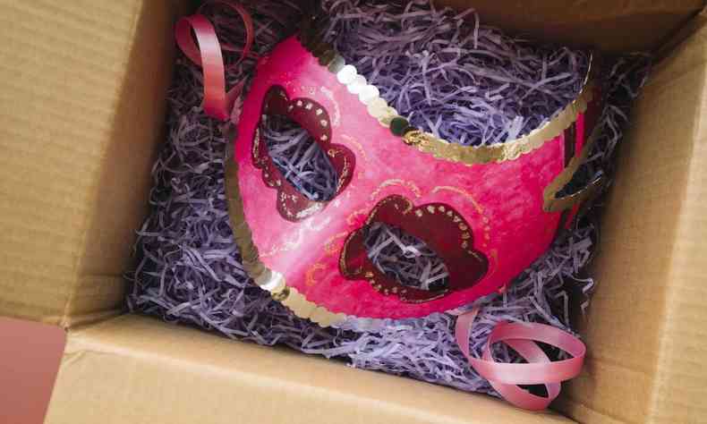 Mscara de carnaval rosa e lantejoulas douradas dentro de uma caixa, que est com a tampa aberta