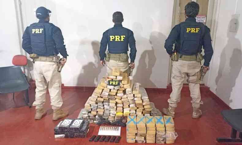 Drogas apreendidas e empilhadas no cho em uma sala. Trs policiais aparecem ao fundo de costas, onde tem a sigla PRF na jaqueta deles. 