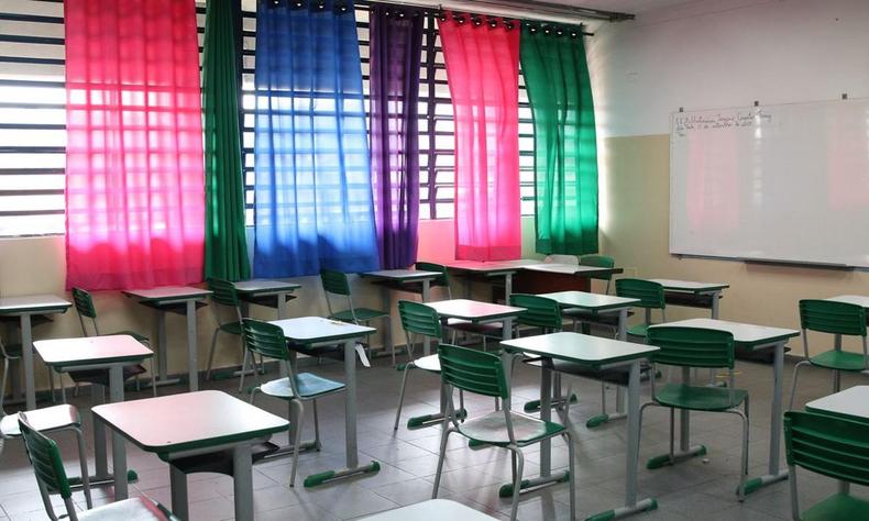 Sala de aula com carteiras vazias. Aparece parte do quadro. Ao fundo h janelas com cortinas coloridas fechadas. 