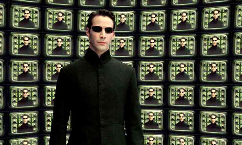 Keanu Reeves veste preto e usa culos escuros em cena de Matrix na qual sua imagem  repetida diversas vezes num painel