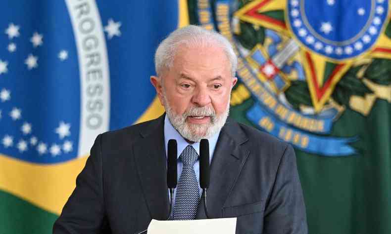 Presidente Lula de terno escuro, camisa azul clara e gravata azul escuro, em plpito com a bandeira do Brasil ao fundo