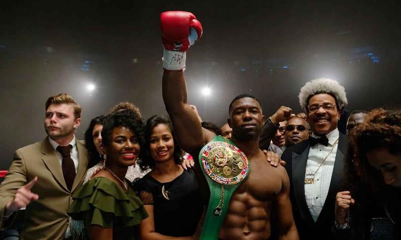Trevante Rhodes faz o papel do lutador Mike Tyson(foto: Star+/divulgação) Ator Trevante Rhodes, como Mike Tyson, levanta o braço esquerdo, cercado de mulheres e de seu empresário