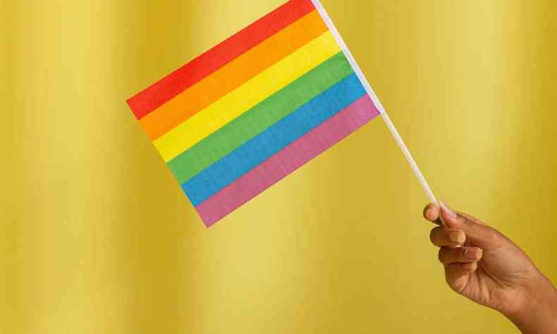 Mo de uma pessoa negra segurando uma bandeira com as cores do arco-iris em um fundo amarelo