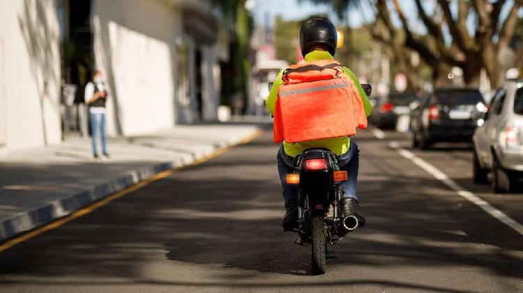 entregador dirige, em imagem que aparece de costas, moto em avenida