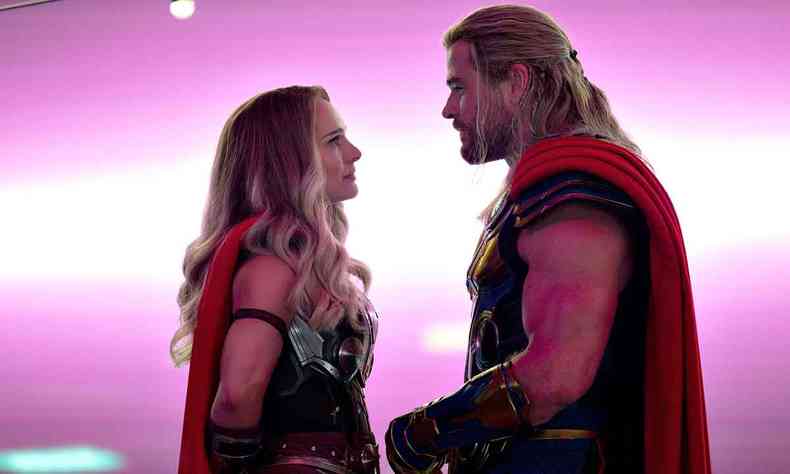 Os atores Natalie Portman e Chris Hemsworth se encaram vestidos como Lady Thor e Thor, sob fundo rosa em cena de thor: amor e trovão 