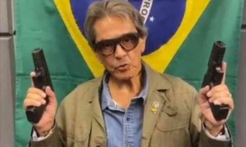 Roberto Jefferson segura uma arma em cada mo e ao fundo tema bandeira do Brasil 