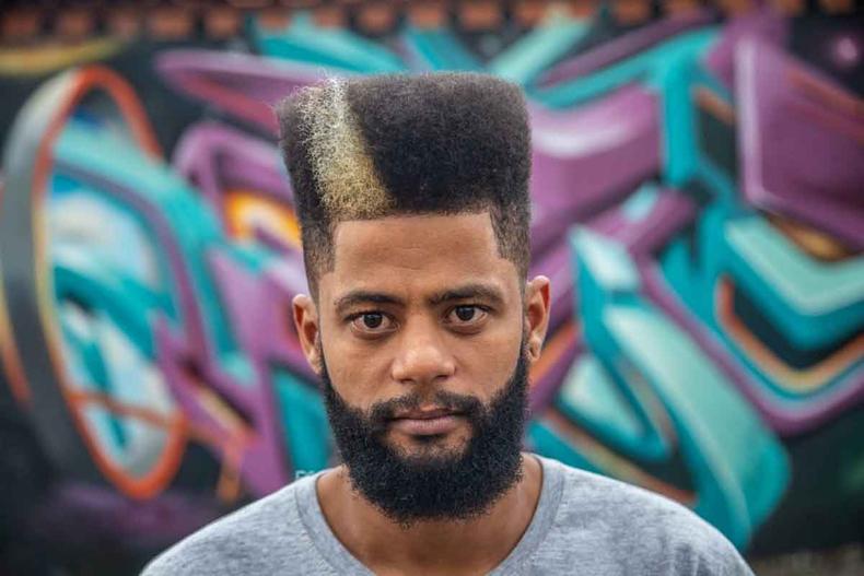 O grafiteiro Leds 2 olha para a cmera, usando corte de cabelo moderno no estilo black power, tendo ao fundo parede pintada em vrias cores