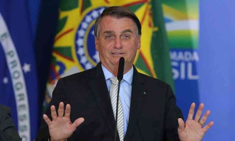 Jair Messias Bolsonaro com as mãos levantadas enquanto fala ao microfone. Ele usa um terno preto com uma camisa azul clara e uma gravata bege. Atrás, vê-se a bandeira do Brasil.