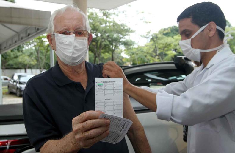 Senhor idoso segura carto de vacina, apontado para a cmera, enquanto recebe vacina de um enfermeiro 