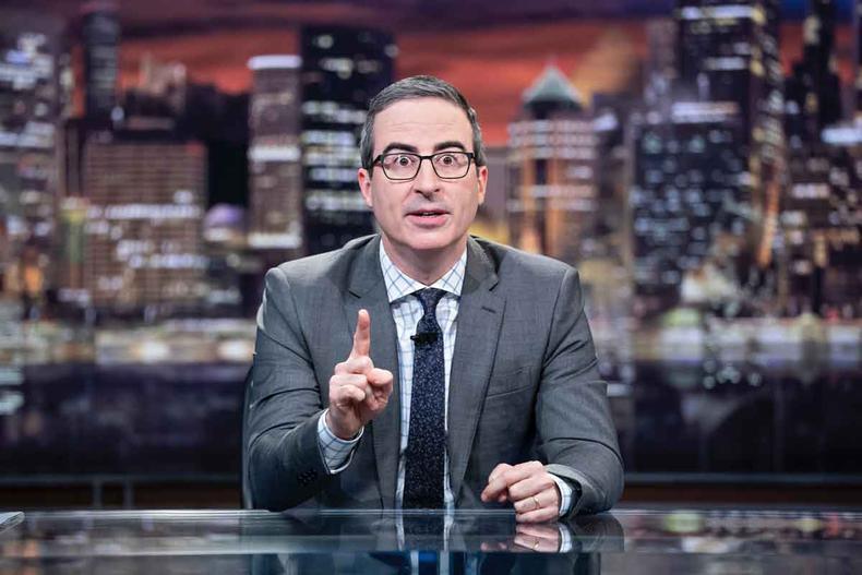 de terno cinza, gravata preta e culos, John Oliver gesticula sentado  mesa que serve de cenrio para o seu talk show