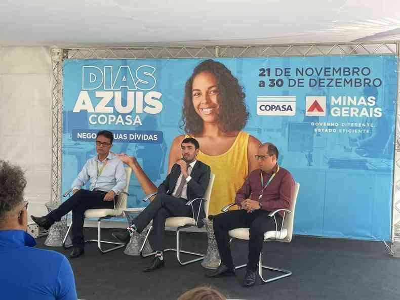 Entrevista coletiva, trs homens sentados e um painel atrs