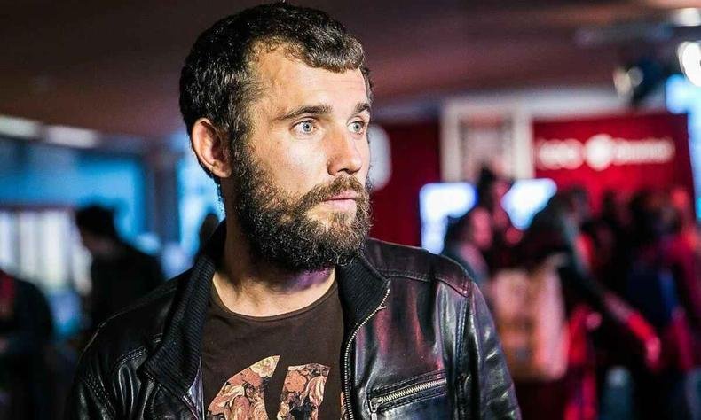 cineasta Mantas Kvedaravicius olha para o lado. ao fundo veem-se letreiros coloridos de lojas