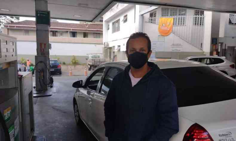 Na foto, o motorista Ernane Costa. O homem está em um posto de gasolina, em frente ao seu carro. Ele usa máscara, um boné e uma jaqueta na cor azul 