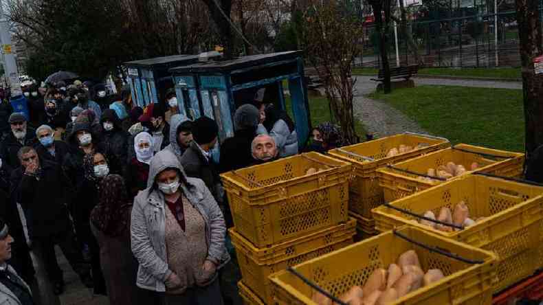 Pessoas agasalhadas e com máscaras em área externa, perto de cestos com pães