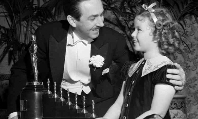 Walt Disney olhando para Shirley Temple diante do uma estatueta do Oscar em tamanho normal e sete miniaturas enfieiradas em uma escada