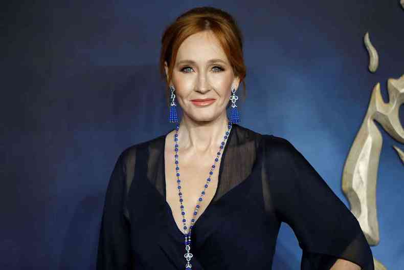 Usando vestido preto, colar e brincos azuis, J.K. Rowling olha para a cmera