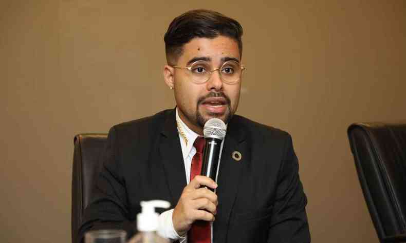 Gregory Rodrigues Roque de Souza, usando terno preto, camisa branca e gravata vermelha, sentado em uma mesa e falando ao microfone