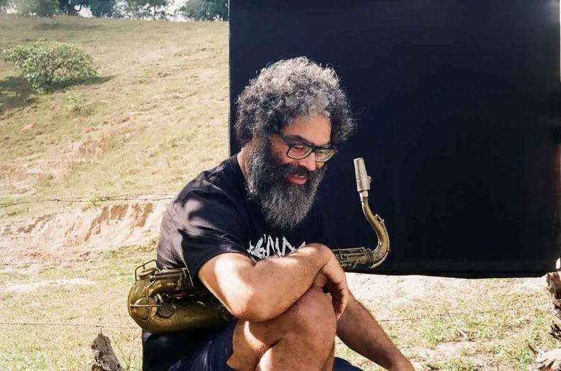 de culos, camiseta e bermuda pretas, agachado num espao ao ar livre segurando seu instrumento musical, Thelmo Cristovam sorri