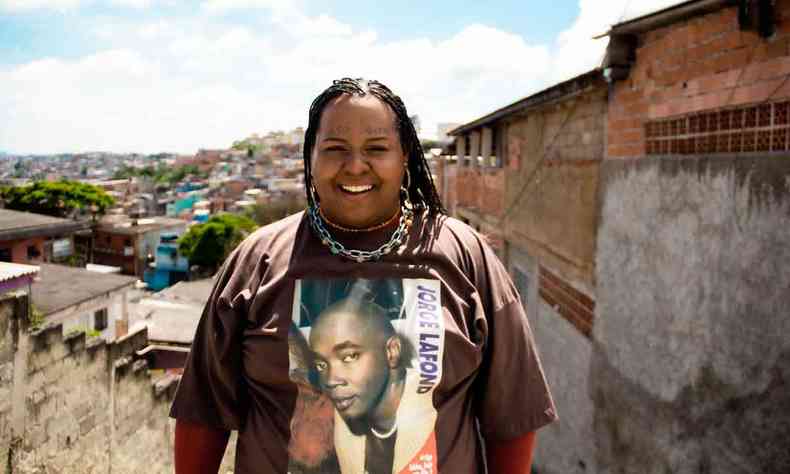 Cantora Jup do Bairro sorri, usando camiseta com o rosto de Jorge Lafond, tendo ao fundo um bairro de periferia e o céu azul