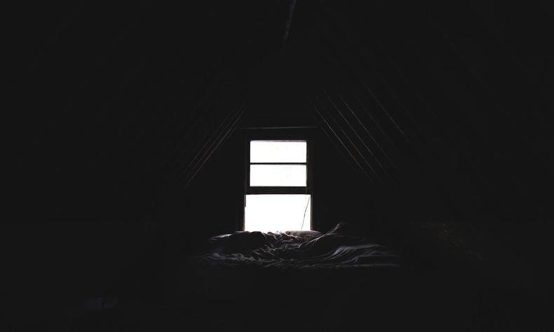 Quarto escuro com cama desarrumada e janela ao fundo