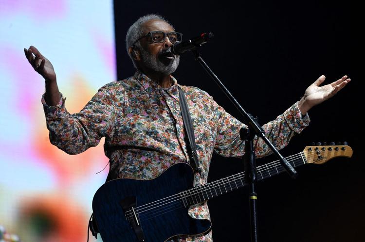 Gilberto Gil cantando de braos abertos em show, com camisa estampada floral