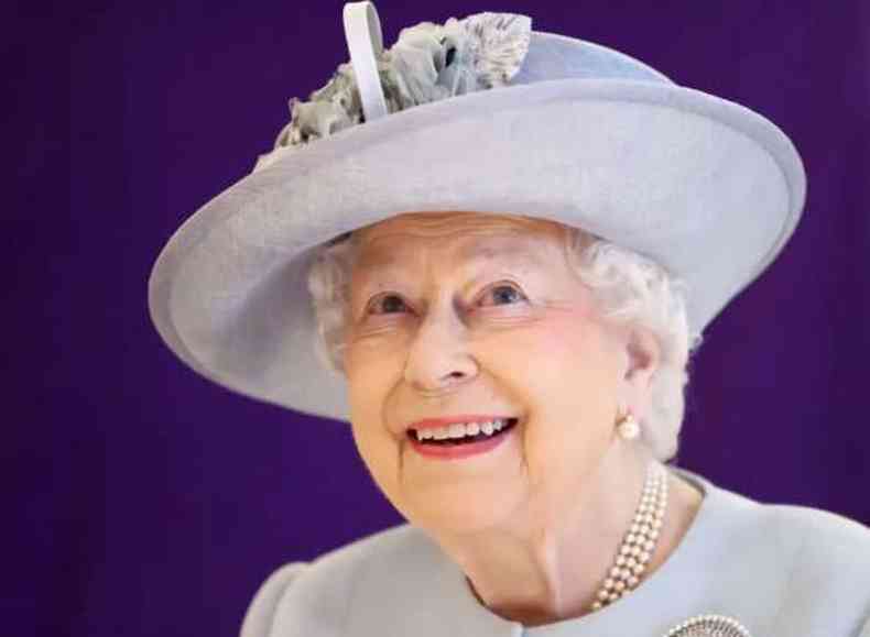 A rainha Elizabeth 2. Ela sorri, olha para cima. A monarca usa um chape lils ornamentado por uma flor e uma blusa de mesma cor, alm de brincos e um colar de prolas. A foto a enquadra dos ombros para cima. 