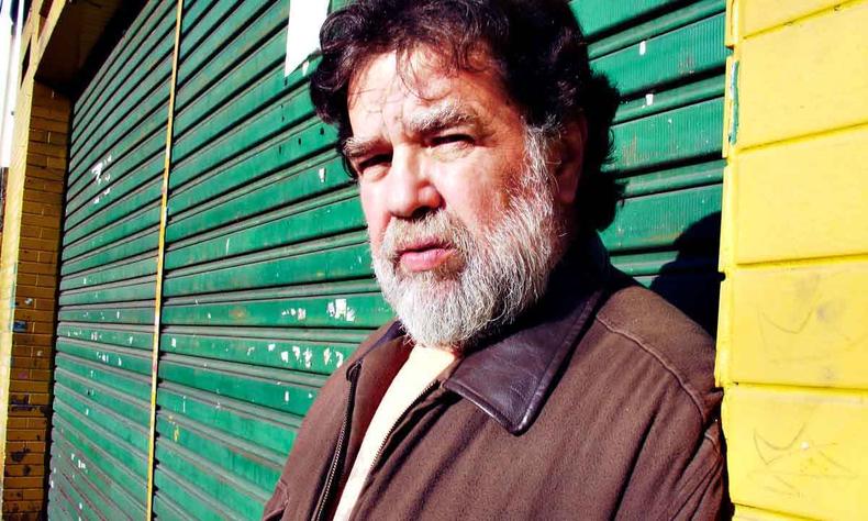 O jornalista Toninho Vaz olha para a cmera, sob o sol, encostado em porta pintada de verde e amarelo 