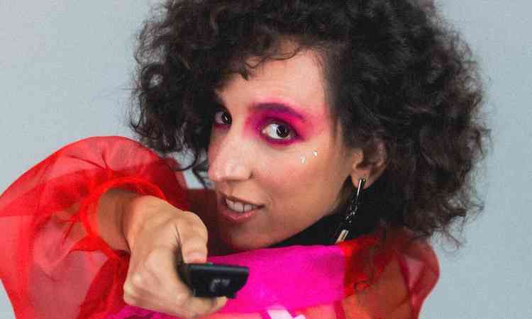 Marina Melo com roupas coloridas e maquiagem, aponta controle remoto para a cmera

