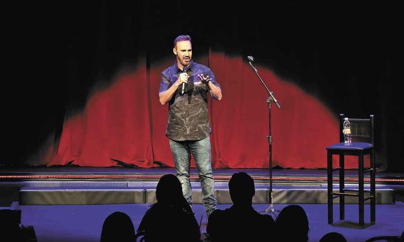 Humorista Diogo Portugal est em cena no palco, segurando o microfone, em frente a plateia 
