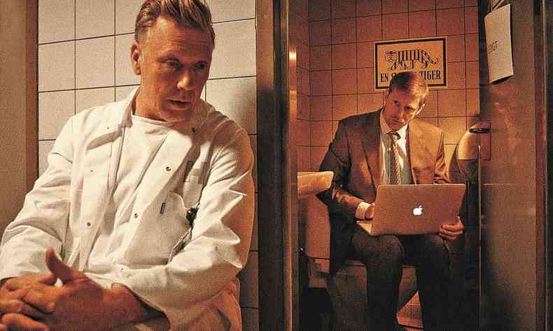 Homem de terno digita em laptop, enquanto outro homem, vestido de camisa branca, do lado de fora da sala, se concentra em ouvir
