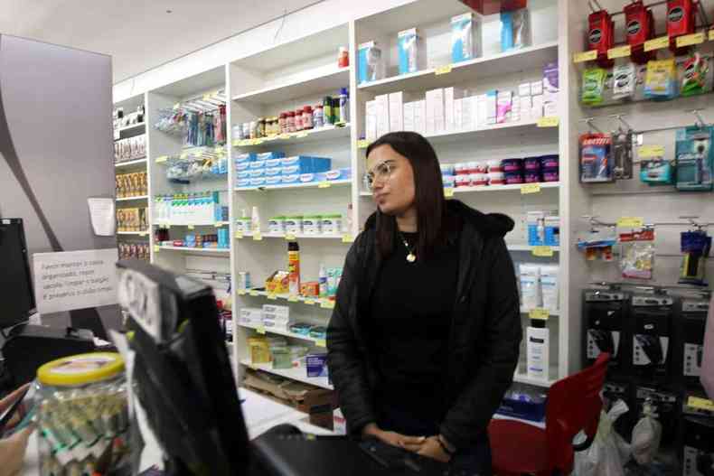 Mulher de pele branca de cabelos pretos longos, usando culos, vestindo roupas tambm pretas. Ao fundo, uma farmcia.