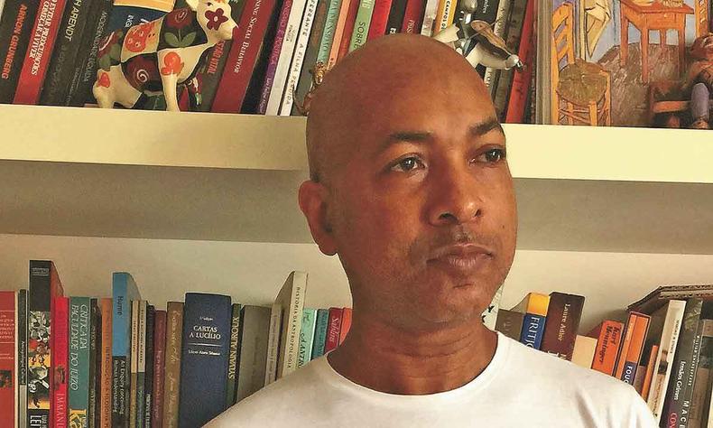 Escritor Marcelo Ariel olha para o lado, tendo ao fundo estante cheia de livros 