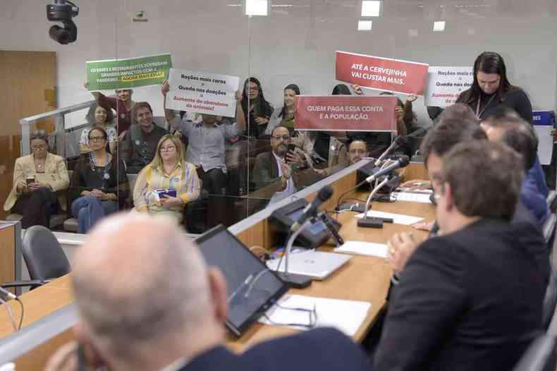 Na sala de reunio da ALMG, pessoas seguram cartazes em protesto contra aumento de tributos proposto pelo governo Zema