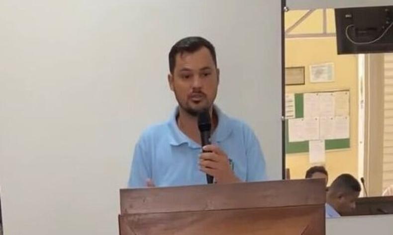 Evandro  um homem pardo de cabelos castanhos lisos. Ele usa uma camisa social azul claro e segura um microfone em frente a um palanque