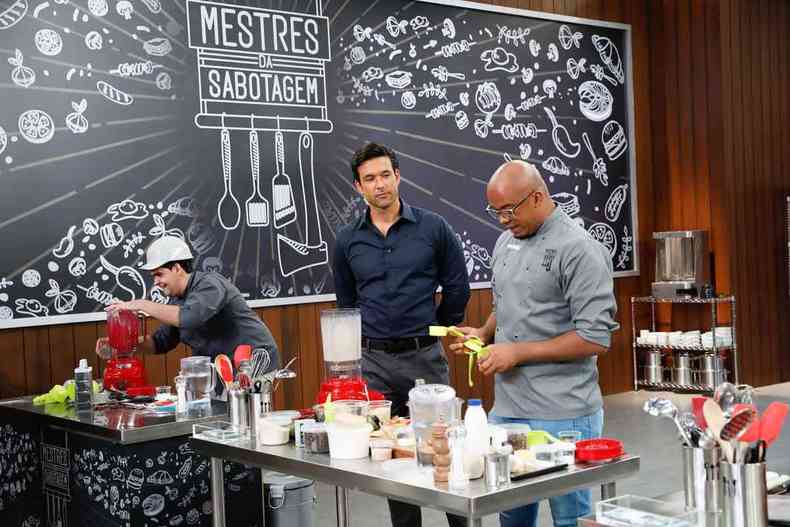 Sergio Marone observa, numa cozinha, dois participantes do programa Mestres da Sabotagem