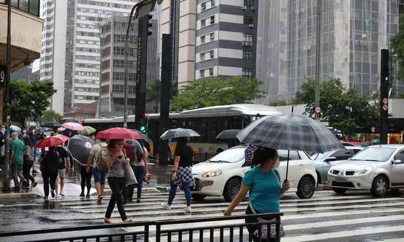 Pessoas de guarda-chuva atravessam uma avenida com carros e nibus