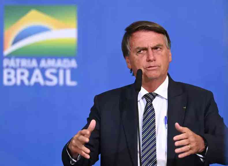 Bolsonaro gesticula com as mãos diante de microfone com fundo azul e bandeira do Brasil 