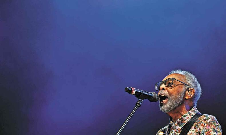 Vestindo casaco com estampa floral, Gilberto Gil canta ao microfone, em palco com fundo azul 