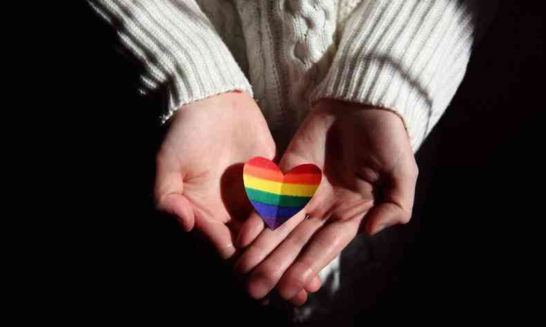 Mãos segurando um coração de papel pintado com as cores do arco-íris 