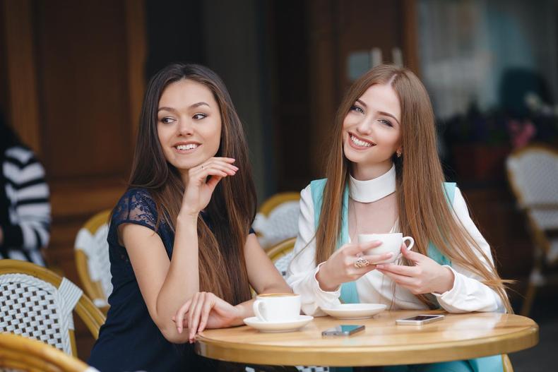 Duas moças sentadas à mesa tomam café e sorriem
