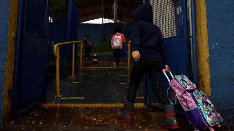 Menina de costas carrega mochila de rodinha, entrando na escola