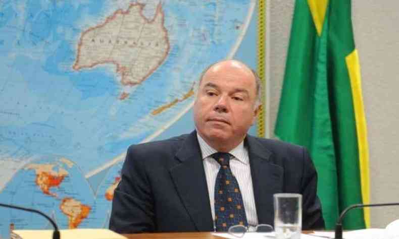 Mauro Vieira sentado  mesa; ao fundo, uma mapa mundi e a bandeira do Brasil