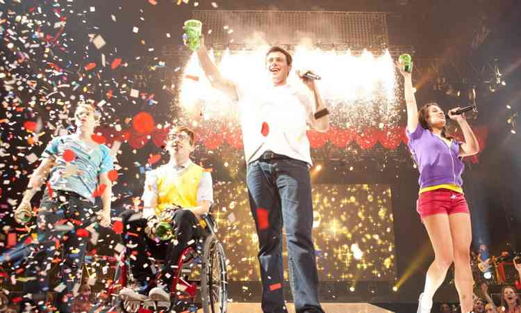 Ator Cory Monteith sorri, segurando o microfone no palco, em cena de Glee
